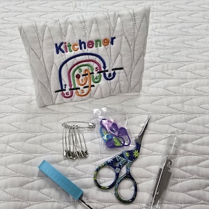 May include: A white, quilted fabric pouch embroidered with the word "Kitchener" and a colorful diagram. The image also shows a set of safety pins, stitch markers, floral scissors, a crochet hook, and a needle threader.