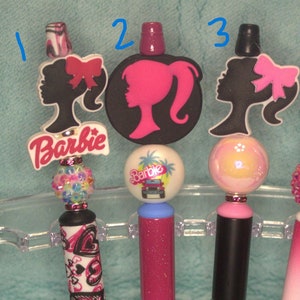 May include: A selection of decorative pens with Barbie-themed designs. The pens feature Barbie silhouettes, bows, and the Barbie logo. The pen barrels are decorated with patterns and glitter. The pens are held in a clear plastic holder.