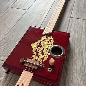 May include: A handcrafted cigar box guitar with a deep red body and a light wood neck. The body features gold lettering and a key-shaped detail. The instrument has three strings and a metal sound hole.