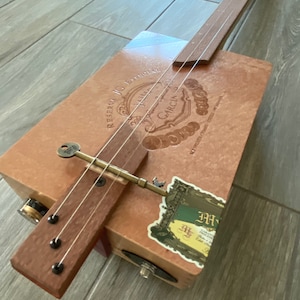 Puede incluir: Una guitarra de caja de puros con un mástil de madera y cuerdas. La guitarra está hecha de una caja de puros reutilizada con una etiqueta que dice "Reserva J.C. Especial", "Alma", "Garcia", y "Hecho a Mano en Cuba".