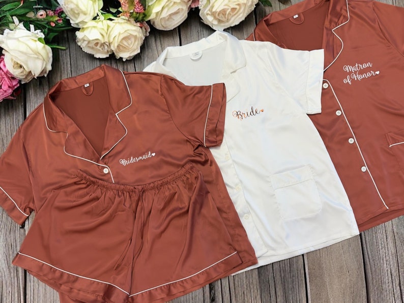 May include: Three satin pajamas with embroidered text. The first pajama set is rust colored with white piping and the text "Bridemaid". The second pajama set is white with white piping and the text "Bride". The third pajama set is rust colored with white piping and the text "Matron of Honor".