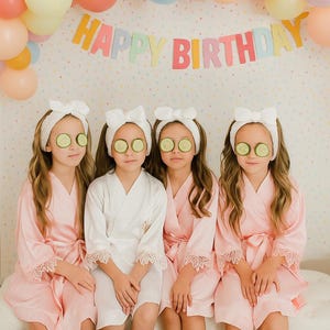 May include: Four young people wearing white headbands with bows and cucumber slices over their eyes, and pink robes, are seated in front of a colorful balloon arch and a banner that reads "HAPPY BIRTHDAY". A table with nail polish and candles is in front.