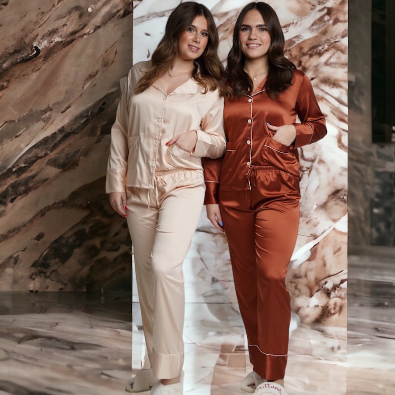 May include: Two women wearing matching satin pyjamas. One woman is wearing a light peach pyjama set and the other is wearing a rust coloured pyjama set. Both sets have a button-up top and long trousers.