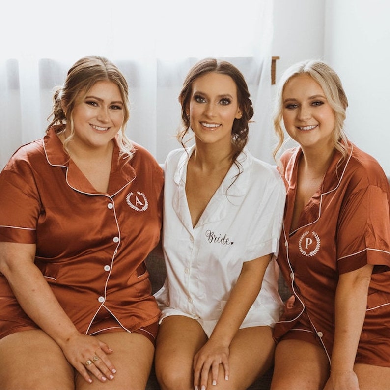 May include: Three women wearing matching pajamas. The woman in the middle is wearing a white pajama set with the word "Bride" embroidered on the front. The women on either side are wearing rust-colored pajama sets with embroidered initials on the front.