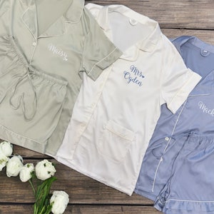 May include: Three sets of pajamas in different colors: sage green, white, and light blue. Each set has a short-sleeved top with a button-up collar and matching shorts. The tops have personalized embroidery with the names "Maisy", "Mrs. Ogden", and "Mack".