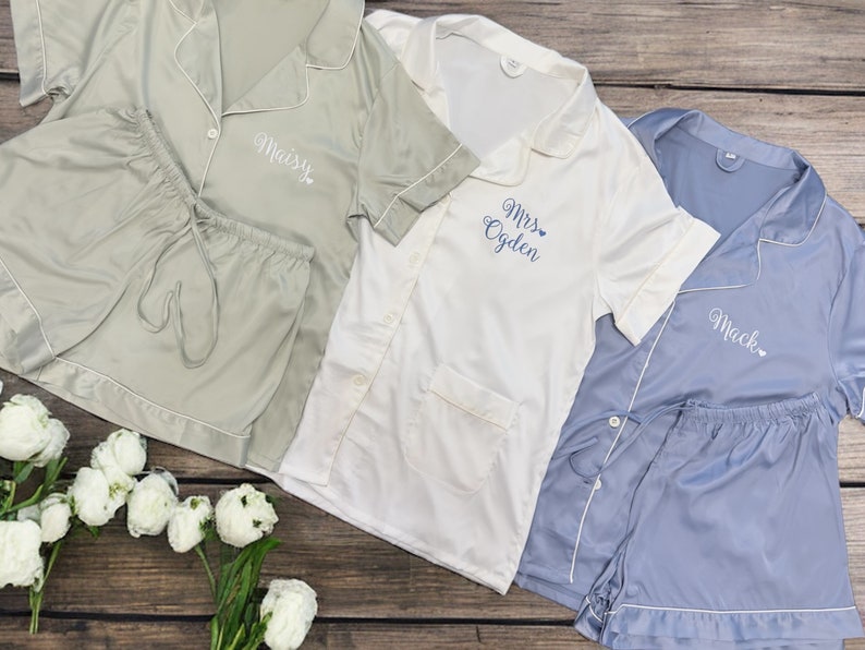 May include: Three sets of pajamas in different colors: sage green, white, and light blue. Each set has a short-sleeved top with a button-up collar and matching shorts. The tops have personalized embroidery with the names "Maisy", "Mrs. Ogden", and "Mack".