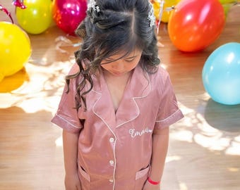 May include: A young girl wearing a pink satin pajama top with white piping and the name "Emma" embroidered on the chest. The girl is standing in a room with colorful balloons in the background.