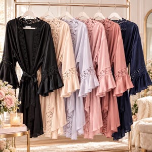 May include: A collection of six satin robes hanging on a gold rack, each featuring lace trim along the neckline and sleeves. The robes are available in black, champagne, light purple, dusty rose, mauve, and navy blue, all with matching waist ties.