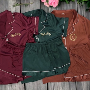 May include: Three sets of matching pajamas in burgundy, forest green, and brown. Each set features a short-sleeved top with a button-up collar and matching shorts. The tops are personalized with gold embroidery, one with the name "Bailey", one with the name "Ashley", and one with a monogram "L" surrounded by a wreath.