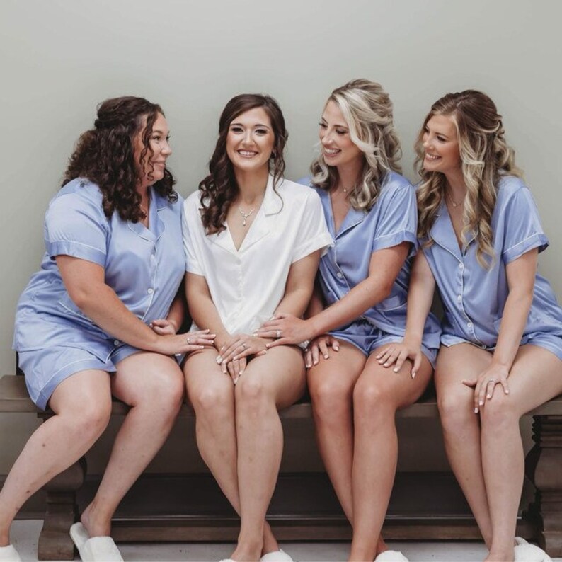 May include: Four women wearing matching blue satin pajamas with white trim. The woman in the center is wearing a white satin pajama top. They are all sitting on a wooden bench and smiling.