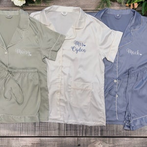 May include: Three sets of matching pyjamas in green, white and blue. The pyjamas have short sleeves and shorts. The green pyjamas have the name "Maisy" embroidered on the front. The white pyjamas have the text "Mrs. Ogden" embroidered on the front. The blue pyjamas have the name "Mack" embroidered on the front.