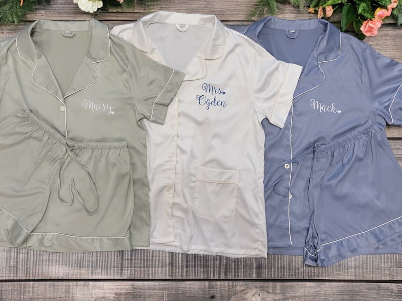 May include: Three sets of matching pyjamas in green, white and blue. The pyjamas have short sleeves and shorts. The green pyjamas have the name "Maisy" embroidered on the front. The white pyjamas have the text "Mrs. Ogden" embroidered on the front. The blue pyjamas have the name "Mack" embroidered on the front.