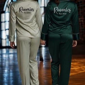 May include: Two people wearing matching satin pajamas, one in a light green color and the other in a dark green. The pajamas have long sleeves and pants with white trim. The text "Roomies - Est 2025" and "Roomies - Est 2023" are printed on the back.