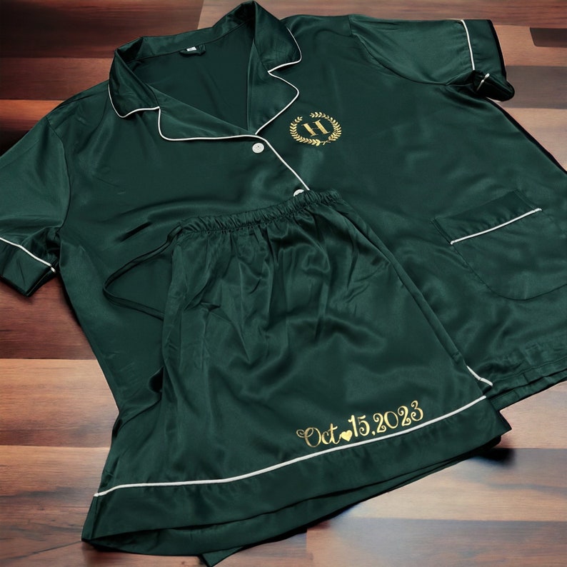 May include: A dark green satin pyjama set with short sleeves and shorts. The top has a white trim and a gold embroidered initial "H" with a laurel wreath. The shorts have a white trim and a gold embroidered date "Oct 15, 2023" with a heart.