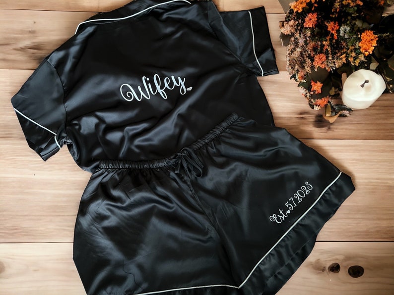 May include: Black satin pyjamas with white piping and embroidery. The top says "Wifey" and the shorts say "Est. 2023".