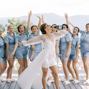 May include: A group of people wearing light blue pajama sets with white piping and white fuzzy slippers. One person in the center wears a white sheer robe with pearl embellishments over a white mini dress and white fuzzy slippers.