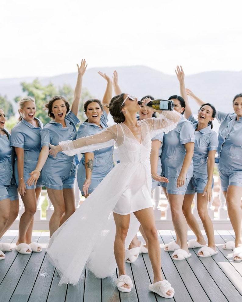 May include: A group of people wearing light blue pajama sets with white piping and white fuzzy slippers. One person in the center wears a white sheer robe with pearl embellishments over a white mini dress and white fuzzy slippers.
