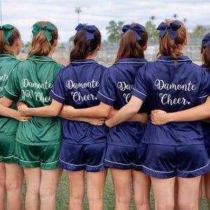 May include: A group of people wearing matching green and navy blue satin pajama sets with white trim. The backs of the shirts have the words "Damonte Cheer" in a cursive font. Each person is wearing a matching bow in their hair.