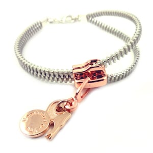 May include: A silver zipper bracelet with a rose gold zipper pull and a rose gold charm with the words "Mastering the Art of You".