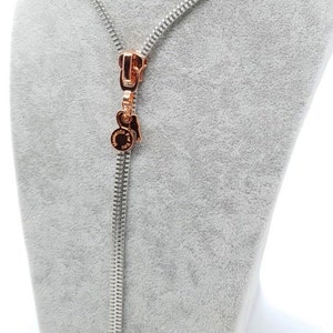 May include: A silver zipper necklace with a rose gold zipper pull and a rose gold charm. The charm has a circular design with the words "Marc Jacobs" inscribed on it.