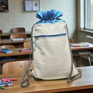 Puede incluir: Una mochila beige con cordón y parte superior con estampado de cachemira azul. La mochila tiene una cremallera azul y correas de cuerda en blanco y negro. La bolsa está sobre un escritorio de madera en un aula.