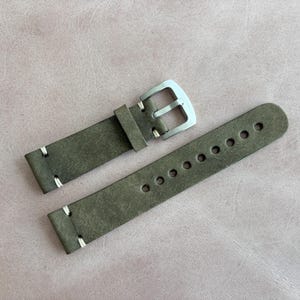 May include: A watch strap made of olive green leather with white stitching. The strap has a silver-colored buckle and multiple adjustment holes. The leather has a distressed appearance.