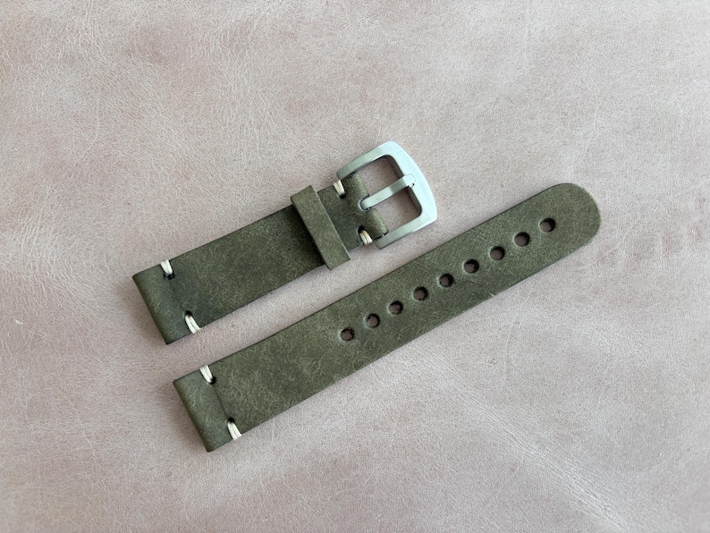 May include: A watch strap made of olive green leather with white stitching. The strap has a silver-colored buckle and multiple adjustment holes. The leather has a distressed appearance.