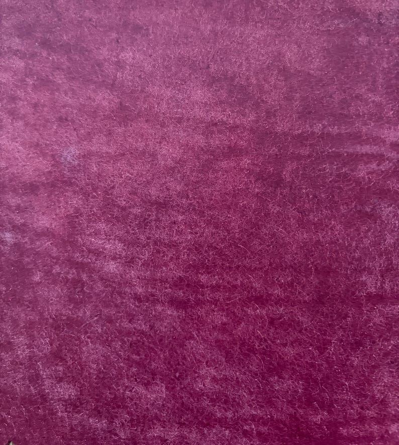 May include: Close-up of a deep magenta fabric with a soft, textured surface. The material has a plush, velvety appearance, with subtle variations in color and light. The fabric's fibers create a rich, tactile feel.