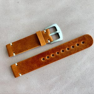May include: A pair of brown leather watch straps with white stitching and a silver-colored buckle. The straps have multiple holes for adjustment and are displayed on a light gray surface.