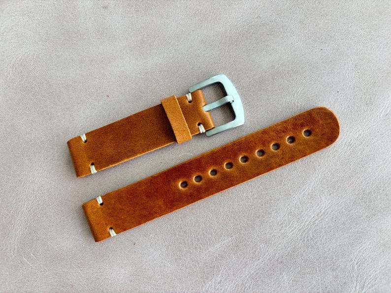 May include: A pair of brown leather watch straps with white stitching and a silver-colored buckle. The straps have multiple holes for adjustment and are displayed on a light gray surface.