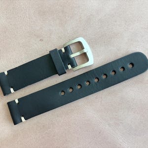May include: Black leather watch strap with white stitching and a silver-colored buckle. The strap has multiple holes for adjustment. The watch band is shown on a light brown surface.