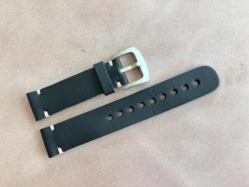 May include: Black leather watch strap with white stitching and a silver-colored buckle. The strap has multiple holes for adjustment. The watch band is shown on a light brown surface.