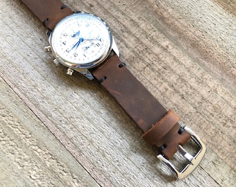 May include: A silver watch with a white face and brown leather band. The watch has a moon phase complication and a date window.