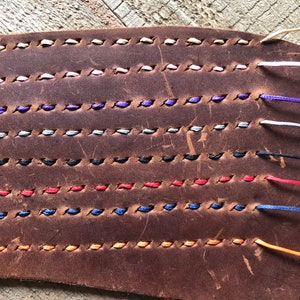May include: Close-up of a brown leather strip with multiple rows of colorful stitching. The stitching is done in a variety of colors, including white, pink, purple, gray, black, red, blue, and orange.