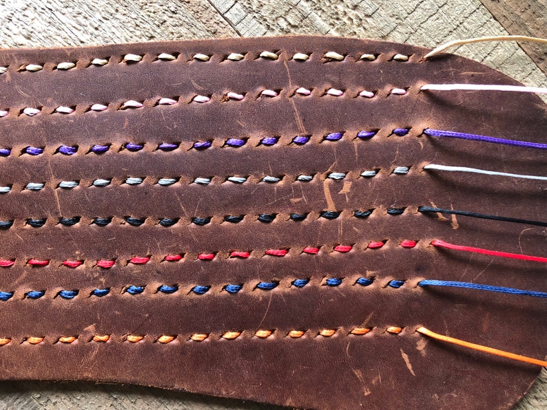 May include: Close-up of a brown leather strip with multiple rows of colorful stitching. The stitching is done in a variety of colors, including white, pink, purple, gray, black, red, blue, and orange.