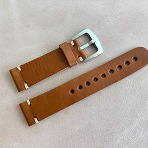 May include: A brown leather watch strap with a silver buckle. The strap has white stitching and multiple holes for adjustment. The watch band is made of a smooth, textured leather.