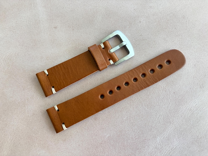 May include: A brown leather watch strap with a silver buckle. The strap has white stitching and multiple holes for adjustment. The watch band is made of a smooth, textured leather.
