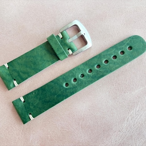 May include: Green leather watch strap with white stitching and a silver buckle. The strap has multiple holes for adjustment. The leather has a textured appearance.