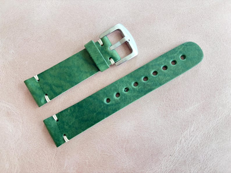May include: Green leather watch strap with white stitching and a silver buckle. The strap has multiple holes for adjustment. The leather has a textured appearance.