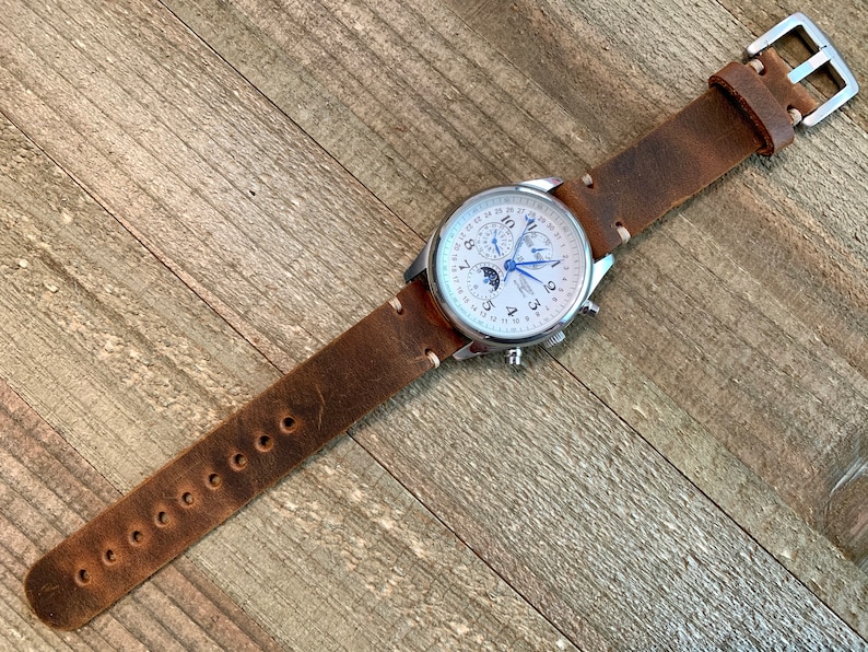 May include: A silver-cased wristwatch with a white face, featuring multiple subdials and a moon phase indicator. The watch has a brown leather strap with white stitching and a silver buckle. The watch is displayed on a wooden surface.