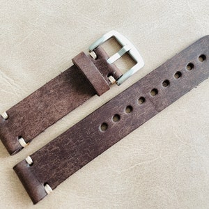 May include: A distressed brown leather watch strap with white stitching and a silver-colored buckle. The strap has multiple holes for adjustment and a rugged, vintage aesthetic. The watch band is a classic accessory.