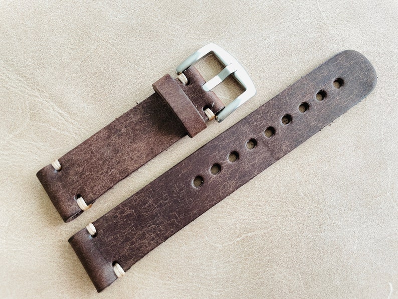 May include: A distressed brown leather watch strap with white stitching and a silver-colored buckle. The strap has multiple holes for adjustment and a rugged, vintage aesthetic. The watch band is a classic accessory.