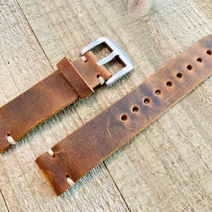 May include: A brown leather watch strap with a silver-colored buckle. The strap has visible stitching and several holes for adjustment. The leather has a distressed appearance, suggesting a vintage or rustic style. The watch band is displayed on a wooden surface.
