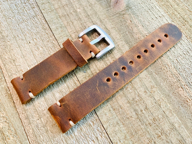 May include: A brown leather watch strap with a silver-colored buckle. The strap has visible stitching and several holes for adjustment. The leather has a distressed appearance, suggesting a vintage or rustic style. The watch band is displayed on a wooden surface.