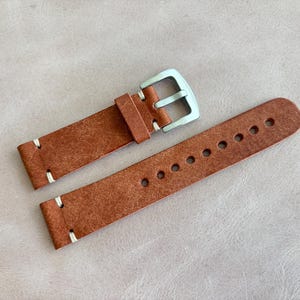 May include: A brown leather watch strap with a silver buckle. The strap has white stitching and multiple holes for adjustment. The leather has a textured appearance.