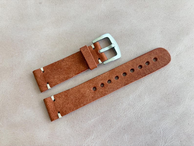 May include: A brown leather watch strap with a silver buckle. The strap has white stitching and multiple holes for adjustment. The leather has a textured appearance.