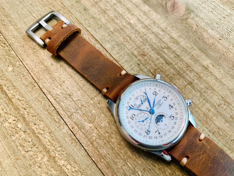 May include: A silver-cased wristwatch with a white face, featuring multiple subdials and a moon phase indicator. The watch has a brown leather strap with white stitching and a silver buckle. The watch is displayed on a wooden surface.