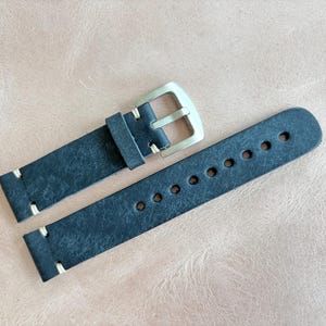 May include: A dark blue leather watch strap with a silver-colored buckle. The strap has white stitching and multiple holes for adjustment. The watch band is shown on a light brown surface.