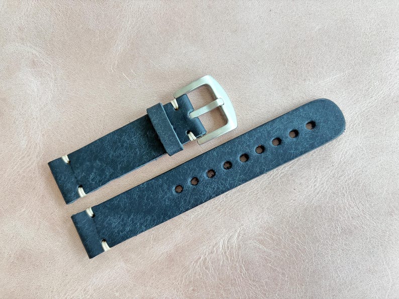 May include: A dark blue leather watch strap with a silver-colored buckle. The strap has white stitching and multiple holes for adjustment. The watch band is shown on a light brown surface.