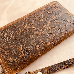 May include: A brown leather wallet with a floral embossed design. The wallet has a zipper closure and a wrist strap.